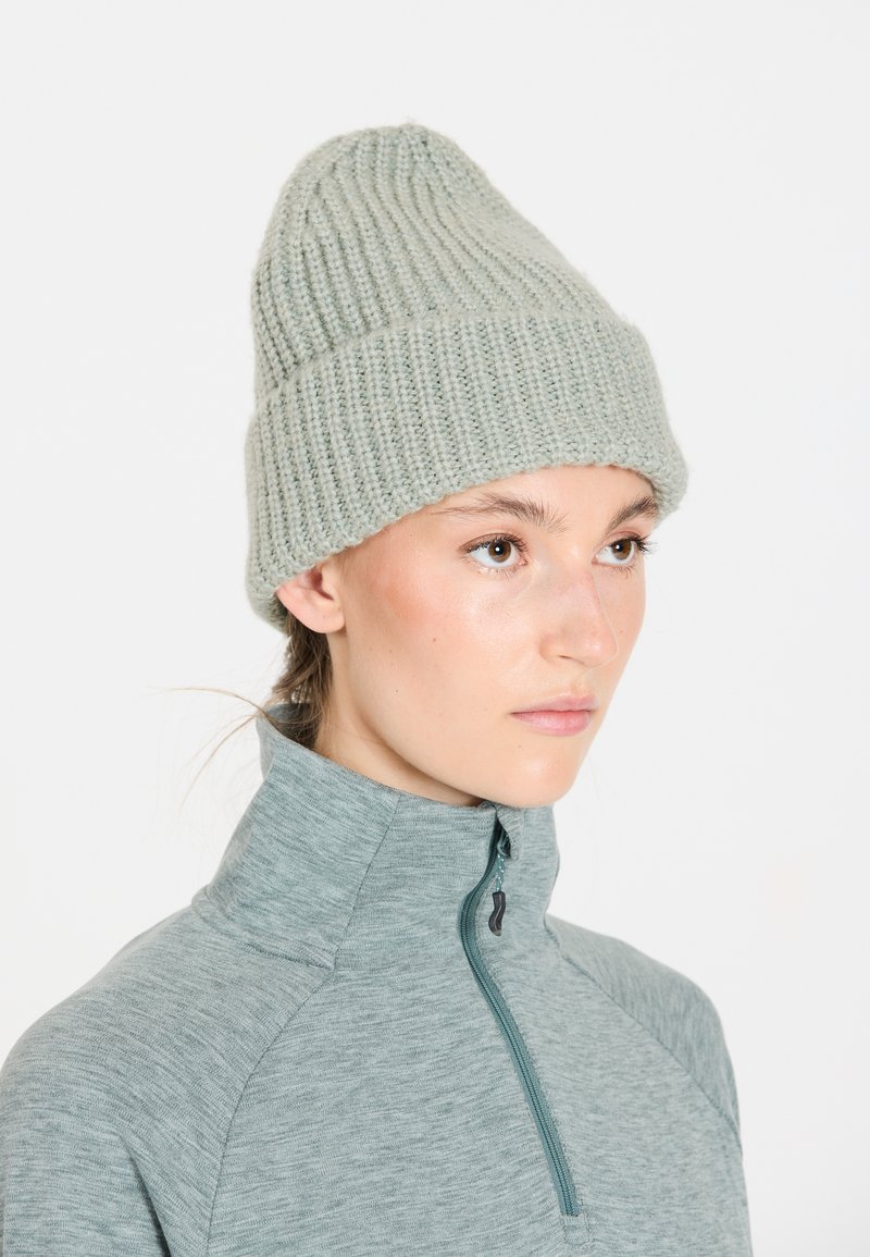 Hellgrübe Strickmütze mit gerippter Textur. Trägt man über einem grauen, hochkragigen, mit Reißverschluss versehenen Sportoberteil. Weiches, wollähnliches Material.