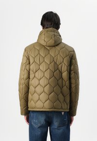 Jachetă puffer matlasată de culoare verde măsliniu, cu guler înalt, având o textură netedă și un model în diamant, purtată peste blugi.