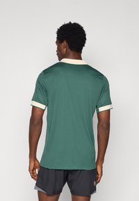 Zöld sporting póló krém színű gallérral és ujjal. Texturált, légáteresztő anyagból készült. A hátsó nézetből a testhez simuló fazon látható.