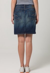 Jeanskjol i mörkblå tvätt, midjelång till mitten av låret, med två bakfickor med böjda sömmar och avslutad fåll, kombinerad med ljusgrå skor.