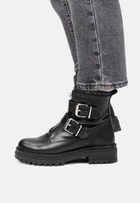 Botas de cuero negras hasta el tobillo con dos hebillas plateadas, una suela resistente y un acabado texturizado, llevadas con pantalones de mezclilla grises.