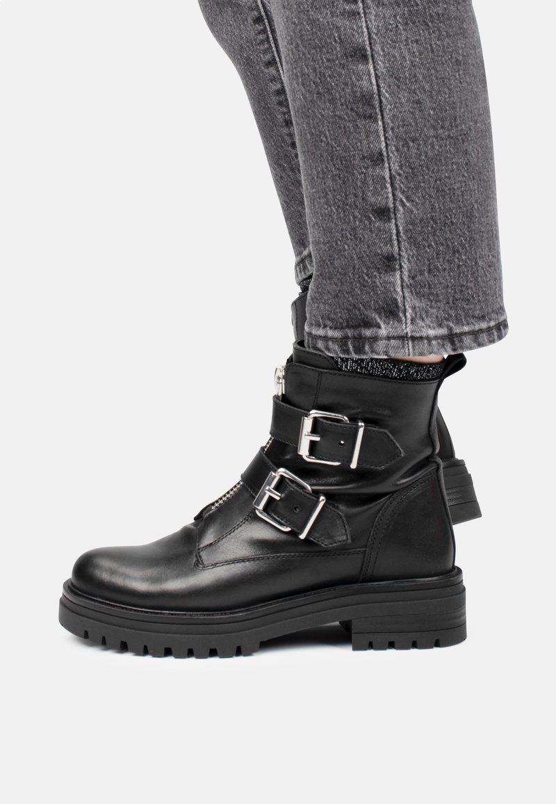 Botas de cuero negras hasta el tobillo con dos hebillas plateadas, una suela resistente y un acabado texturizado, llevadas con pantalones de mezclilla grises.