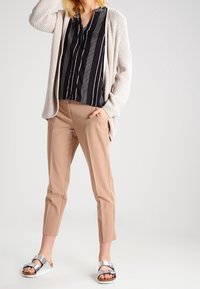 Personne portant un pantalon beige court, un chemisier rayé noir et blanc, un cardigan en maille crème et des sandales argentées, debout devant un fond uni.