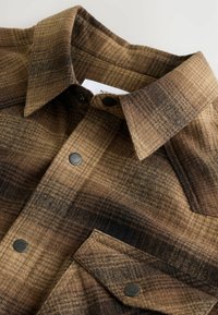 Bruin en zwart geruite shirt met drukknopen en een puntige kraag, toont een close-up van het bovenste deel van de borst en het zakgebied.