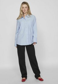 Camisa de botones azul claro con cuello, dos bolsillos en el pecho y mangas largas, combinada con pantalones negros y zapatos planos rojos.