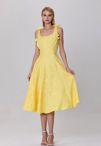 Femme portant une robe midi jaune sans manches avec des nœuds aux épaules, tenant la jupe, se tenant devant un fond gris clair uni.