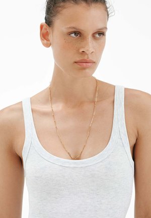 Joven con el cabello recogido, llevando una camiseta sin mangas blanca y un largo collar de cadena dorada, mirando ligeramente hacia un lado.