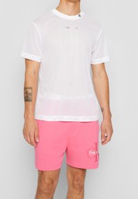 Bílý síťovaný tričko s vroubkovaným kulatým výstřihem a krátkými rukávy; zdobeno barevným textem "To je". Doplněno růžovými šortkami s logem.