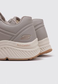 Zapatillas de deporte grises con un acabado texturizado, que cuentan con una suela pronunciada, un collar acolchado y una etiqueta de "Arch Comfort" en naranja claro.