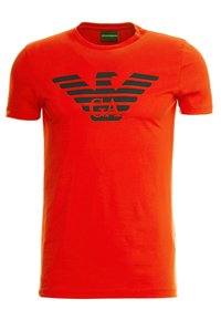 Camiseta de algodón roja con cuello redondo, que presenta un logo de águila negra y acentos a rayas en el pecho. Mangas cortas.