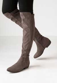 Stivali in suede grigio al ginocchio, con punta affilata, tacco piatto e dettagli con zip laterale, esposti su uno sfondo neutro.