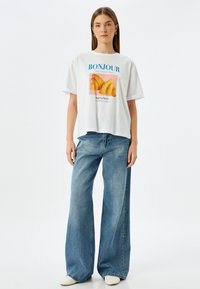 Witte oversized t-shirt met de tekst "BONJOUR" en een croissant graphic. Gecombineerd met wijdvallende blauwe denim jeans en crèmekleurige schoenen.