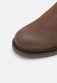 Bota de cuero marrón con punta redonda, suela negra contrastante y un sutil detalle de costura a lo largo del borde. Textura suave, diseño duradero.
