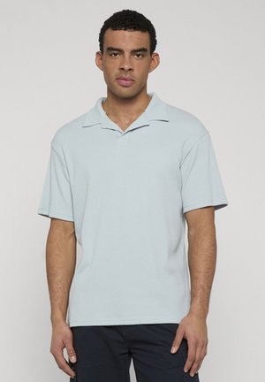 Junger Mann mit kurzen, lockigen Haaren, der ein hellblaues kurzärmeliges Poloshirt und dunkle Hosen vor einem schlichten grauen Hintergrund trägt.