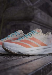 Pair of light beige running shoes with orange stripes and blue laces placed on a wooden surface against a blurred dark background.