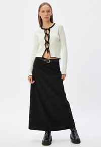 Cárdigan blanco acanalado con detalles negros, con un cierre de encaje en el frente. Combinado con una falda maxi negra y botas gruesas negras.