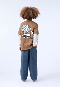 Brun oversized t-skjorte med en tegneseriehjernen på en skateboard; parret med vide, blå denimjeans. Hvite ermer har fargerike grafikk.