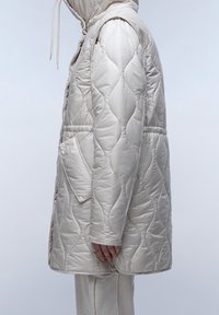 Prošívaná péřová bunda světle béžové barvy, vybavená kapucí, velkými bočními kapsami a hladkým, lesklým povrchem s jemným vlnkovým vzorem.