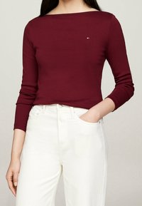 Top lungo bordeaux a maniche lunghe con scollo a barca, realizzato in tessuto morbido, dotato di un piccolo logo sul petto, abbinato a pantaloni bianchi a vita alta.