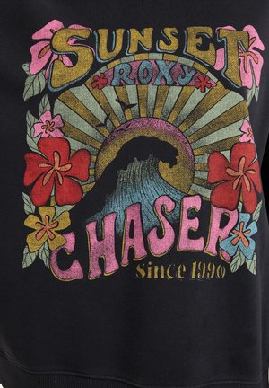 Svart hettegenser med et fargerikt trykk som viser en bølge, blomster, og teksten "SUNSET ROXY CHASER siden 1990." Teksten er fet og variert i farge.