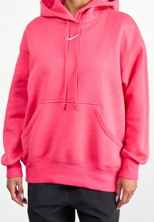 Jasnoróżowa, oversize'owa bluza z kapturem, z przednią kieszenią i sznurkami przy kapturze, noszona z czarnymi spodniami, z małym białym logo Swoosh na piersi.