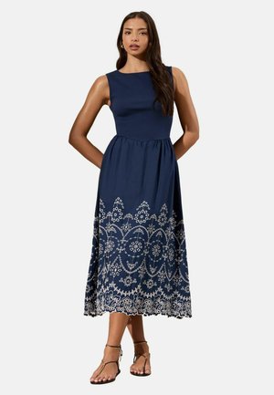 Žena nosi bezrukávové midi šaty námorníckej modrej farby s bielou kvetinovou výšivkou na sukni, kombinované s čiernymi plochými sandálmi s remienkami, stojaca pred bielym pozadím.