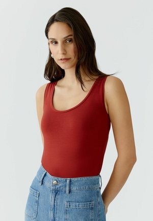 Vrouw met lang donker haar, een mouwloze rode top ingebouwd in een high-waisted blauwe jeans, staande tegen een effen lichte achtergrond.