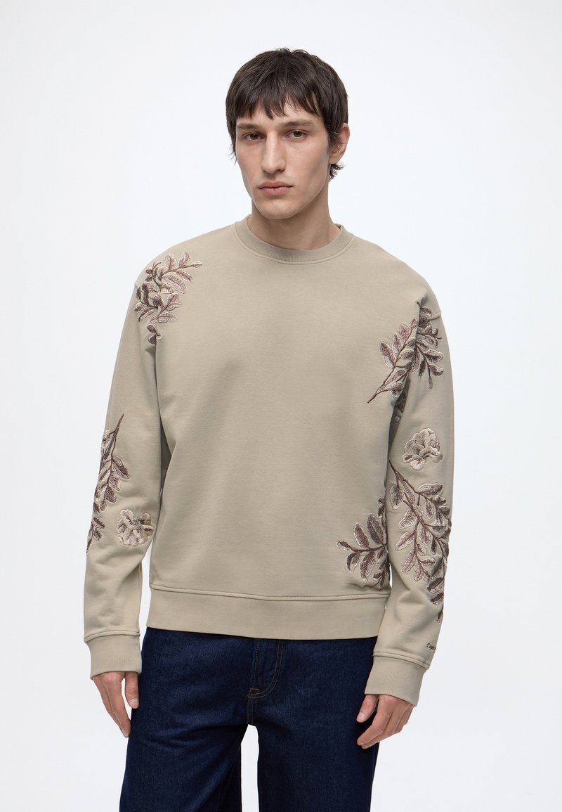 Homme portant un sweat-shirt beige avec une broderie florale marron sur les manches et la poitrine, associé à un jean bleu foncé, debout devant un fond blanc.