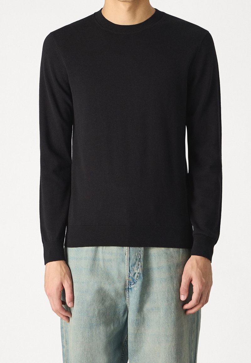 Homme portant un pull noir uni à manches longues avec un col rond et un jean en denim bleu clair, debout devant un fond blanc.