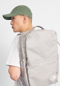 Jauns cilvēks zaļā cepurē, kurš valkā lielu gaiši pelēku North Face mugursomu pār balto kreklu, skatās pa kreisi uz vienkrāsaina fona.