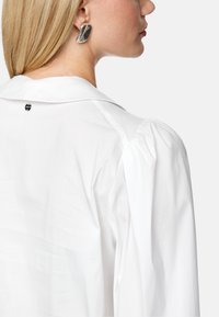 White blouse with a classic collar, puffed sleeves, and a visible button accent on the back. Model wears a large, curved silver earring.