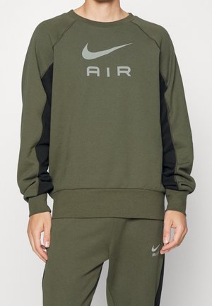 Person, die einen olivgrünen Nike Air Sweatshirt und passende Hosen mit schwarzen Seitenpanelen und grauen Nike-Logos trägt.
