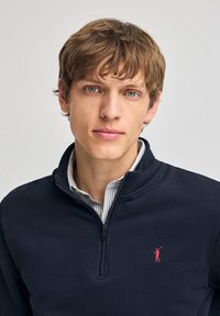 Navy zip-up pullover van zachte stof, met een opstaand kraag en een klein rood logo op de borst, gedragen over een gestreept overhemd met kraag.