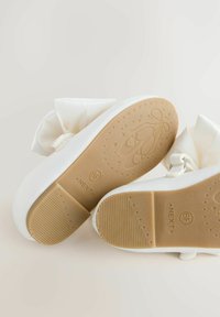 Weiße Kinderschuhe aus Stoff mit einer Schleifendetail, ausgestattet mit einer strukturierten, hellbraunen Gummisohle, die mit einem Sternmuster und Logo geprägt ist.