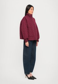 Quilted, maroon puffer jacket with a high collar, side pockets, and ribbed cuffs, paired with loose dark blue jeans and black heeled shoes.