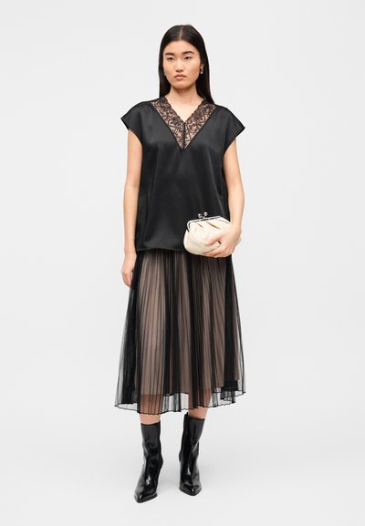 Μαύρο σατέν τοπ με δαντελένιο V-λαιμόκοψη, συνδυασμένο με πλισέ μαύρη και καφέ φούστα και μαύρες μπότες μέχρι το γόνατο, κρατώντας ένα κρεμ φάκελο.