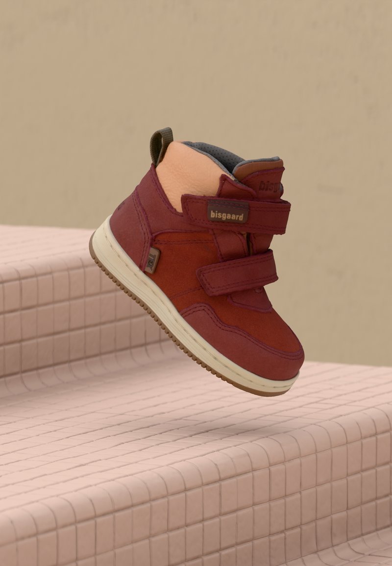 Dětská červená a růžová vysoká teniska se dvěma suchými zipy, béžovou podrážkou a poutkem, vznášející se nad růžovými dlaždicemi.