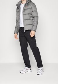 Grijze pufferjas met capuchon, voorzien van horizontale stiksels en een klein logo, gecombineerd met zwarte cargobroeken en witte sportschoenen.