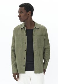 Camisa de botones verde oliva en un tejido ligero, con dos bolsillos en el pecho y un cuello estructurado. Llevada sobre una camiseta negra de canalé.