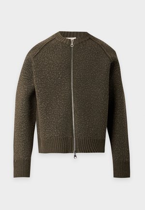 Homme debout portant un pull zippé vert olive foncé, une chemise noire boutonnée, un pantalon ample olive et des chaussures marron foncé avec des lacets rouges.