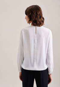 Blusa bianca a maniche lunghe con una leggerissima lucentezza, con dettaglio a chiave sul retro, scollatura rotonda e polsini arricciati. Tessuto liscio, vestibilità ampia.