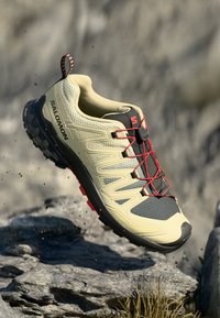 Zapatilla deportiva de color amarillo claro con acentos en negro y rojo, parte superior texturizada, suela elevada y sistema de cordones elásticos. Diseñada para uso exterior.