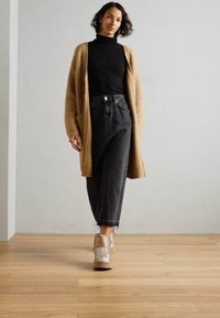Chunky, ribbet beige cardigan, tettsvart høyhalset genser, høy-midje mørke jeans og glitrende gull støvletter med kilehæler, på tregulv.