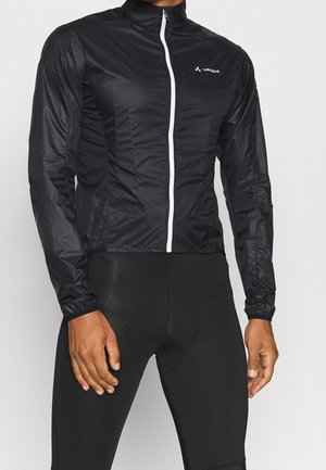 Mann trägt eine schwarze, leichte Jacke mit Reißverschluss und weißem Logo auf der Brust, kombiniert mit schwarzen Radhosen.