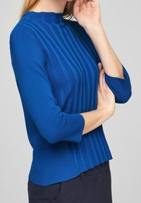 Blårandig stickad topp med hög krage och trekvartsärmar. Texturerade vertikala ränder skapar visuell intresse. Mjuk, figurnära design.