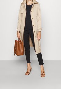 Beige trenchcoat med bälte i midjan, bruna knappar och sidofickor. Svart åtsittande topp och byxor, parat med brun bucket bag och klacksandaler.