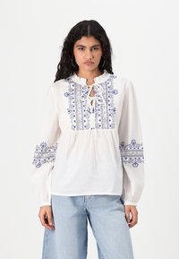 Blouse blanche à manches longues avec un col à volants, ornée d'une broderie florale et géométrique bleue sur le devant et les manches. Coupe ample.