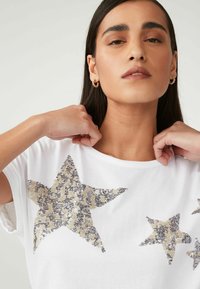 T-shirt blanc avec de multiples formes d'étoiles en sequins argentés et dorés ; manches courtes ; coupe décontractée ; col rond.