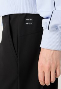 Zwarte sportbroek met een stijlvol ontwerp, voorzien van een zijzak en een logo-patch. Een lichtblauwe opgerolde mouw is zichtbaar in het beeld.