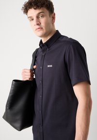 Camicia blu navy a maniche corte in tessuto traspirante con colletto a punta e logo "BOSS" bianco. Il modello tiene in mano una borsa a tracolla in pelle nera.